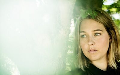 Lyshåret ung kvinde står ved siden af et træ og kigger ud til venstre