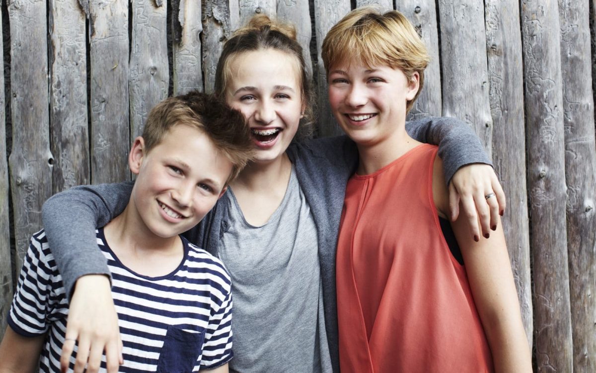 To drenge og en pige griner til kameraet foran et hegn