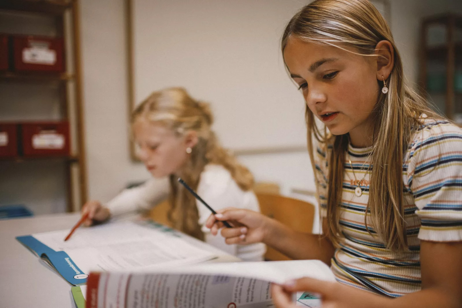 Lyshåret pige læser en skolebog med en anden lyshåret pige i baggrunden