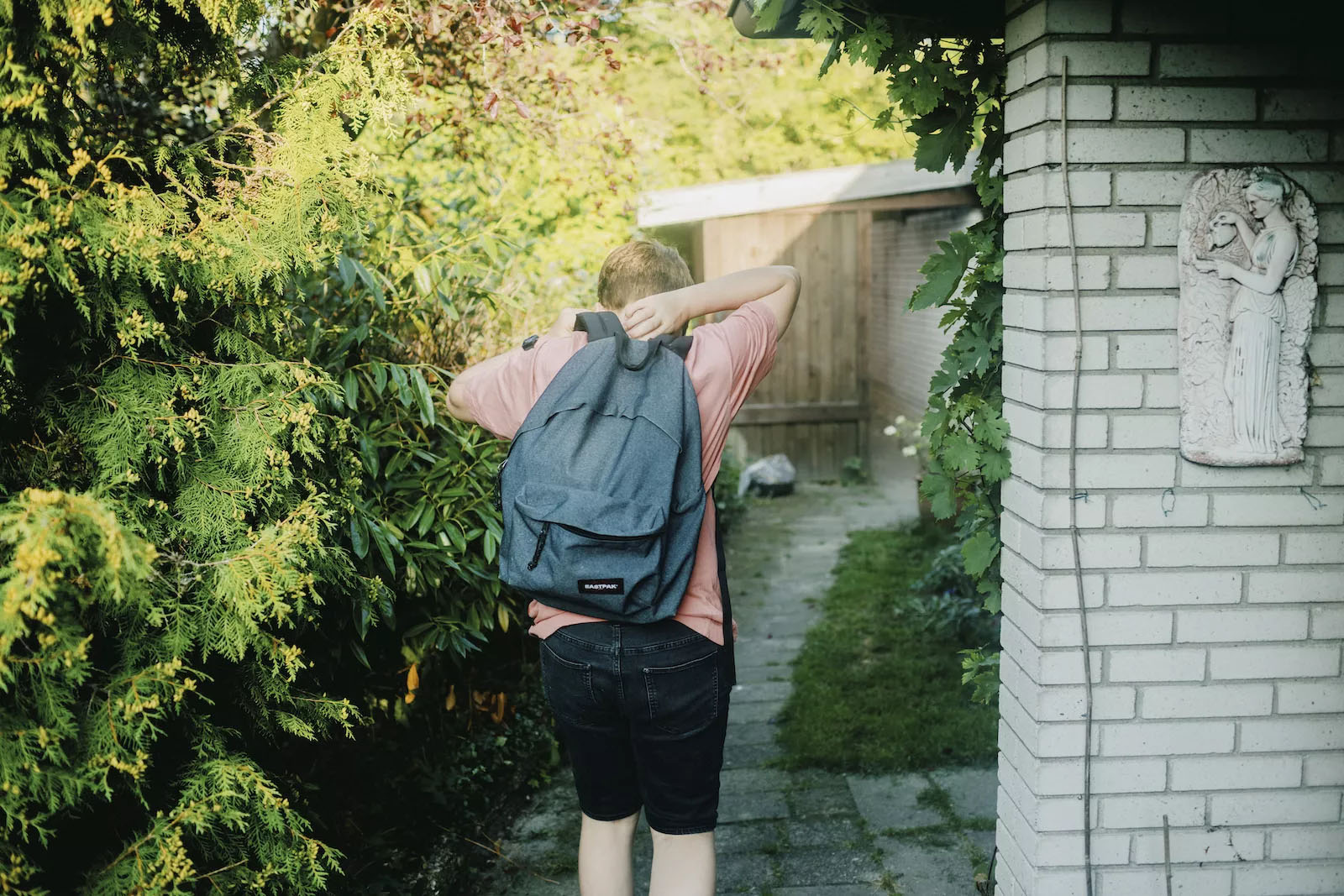 Lyshåret dreng i lyserød trøje, mørke shorts og en blå rygsæk set bagfra